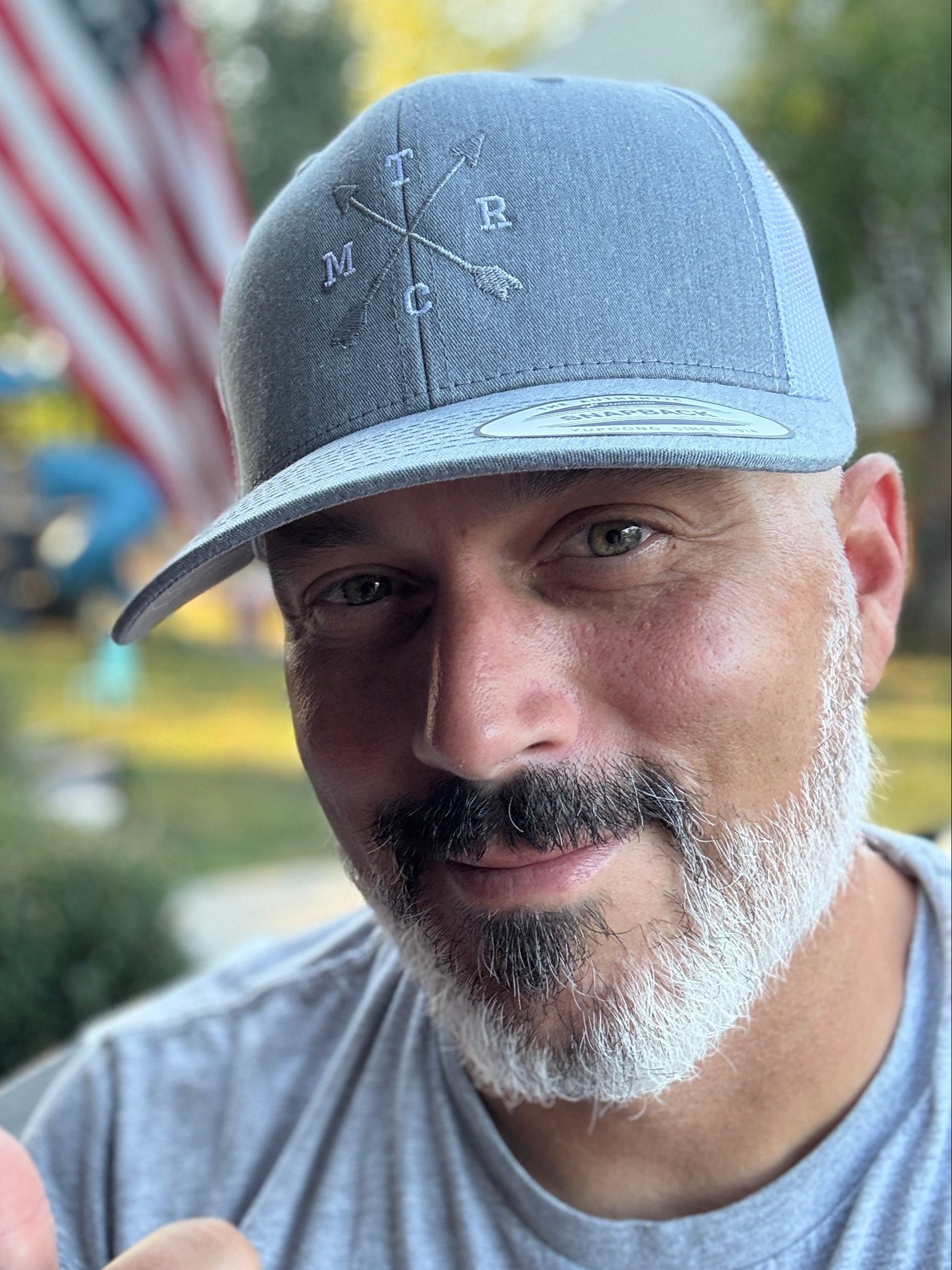 Man wearing a gray cap with a logo, giving a thumbs-up gesture outdoors.